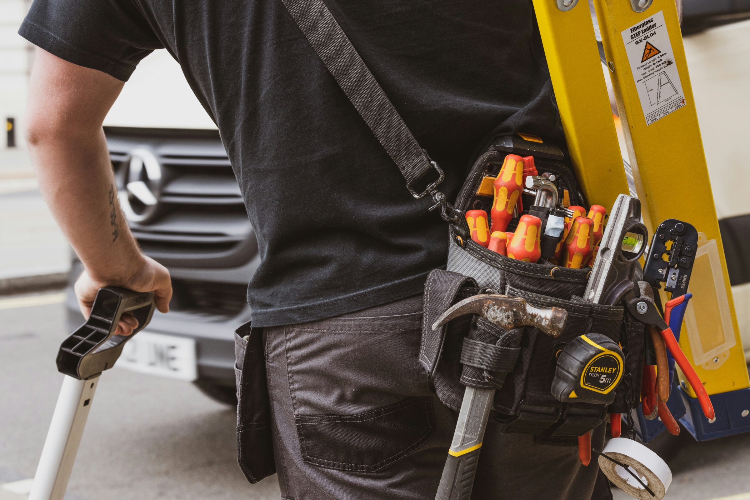 A tradesman with a loaded tool belt and a step ladder, walking toward a job